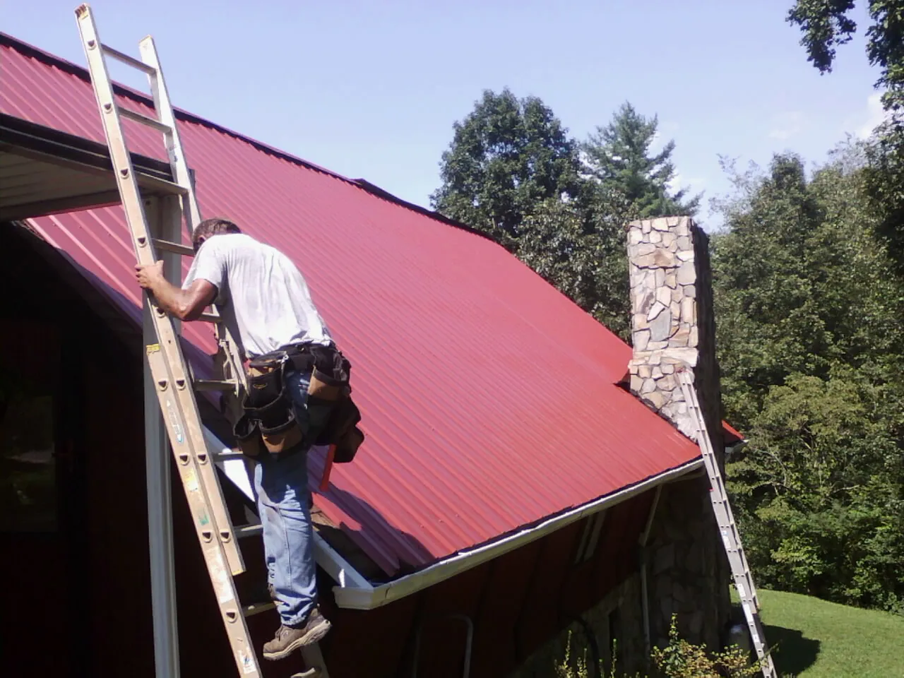 Roofer on a ladder beside a red metal roof and stone chimney — Soffit & Fascia Repair by Roofing Brothers in Rockmart